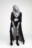 AI-generated fashion portrait of a woman in a long sheer coat, layered necklaces, and boots, striking a confident pose against a blank background